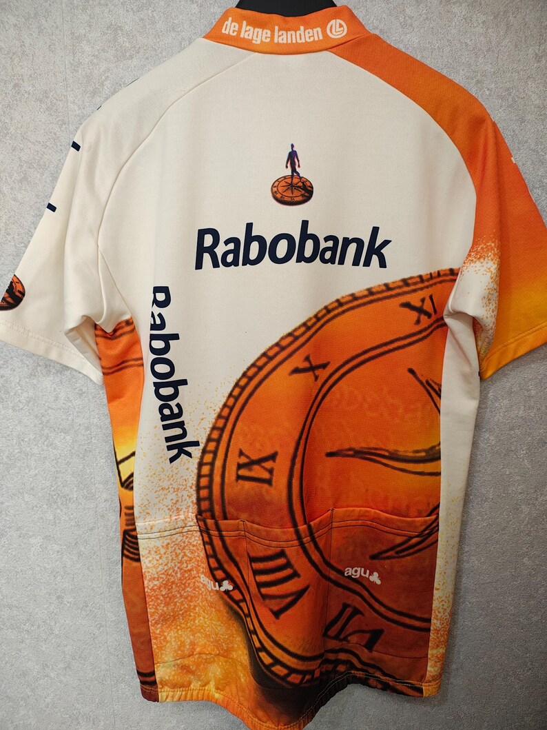 Puede incluir: Maillot de ciclismo color crema con detalles en naranja y el logo "Rabobank". El maillot presenta un gran dise&ntilde;o de reloj y el texto "de lage landen". La espalda tiene tres bolsillos.