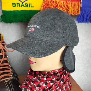 May include: A gray cap with ear flaps and a curved brim, featuring the text "RL JEANS CO." and a small American flag. The cap is displayed on a mannequin head with a red patterned scarf and a black boot.