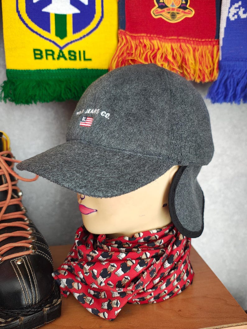May include: A gray cap with ear flaps and a curved brim, featuring the text "RL JEANS CO." and a small American flag. The cap is displayed on a mannequin head with a red patterned scarf and a black boot.