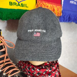 May include: A gray Polo Jeans Co. baseball cap with a curved brim. The cap features white embroidered text and a small American flag emblem. The hat is displayed on a mannequin head, with a red patterned scarf visible below.