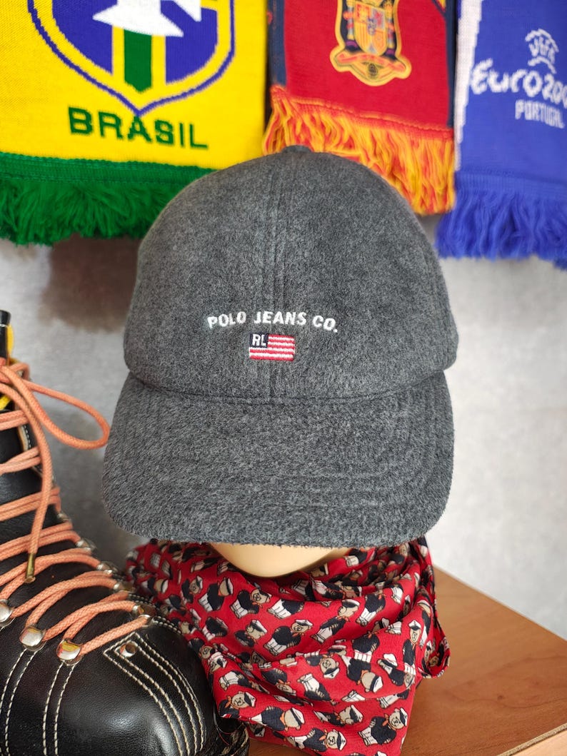 May include: A gray Polo Jeans Co. baseball cap with a curved brim. The cap features white embroidered text and a small American flag emblem. The hat is displayed on a mannequin head, with a red patterned scarf visible below.