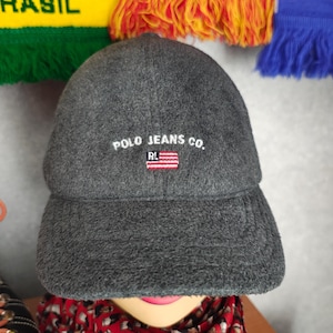Pode incluir: Boné de beisebol cinza, mistura de lã, com aba curva. O boné apresenta o texto "POLO JEANS CO." acima de um pequeno emblema da bandeira americana. Um chapéu de estilo clássico, adequado para uso casual.
