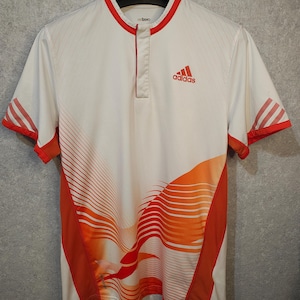 Puede incluir: Camiseta deportiva blanca con detalles en naranja y rojo. Presenta el logo de Adidas en rojo en el pecho. Tiene cuello mandarín y mangas cortas con rayas rojas. El diseño incluye líneas curvas en naranja y rojo.