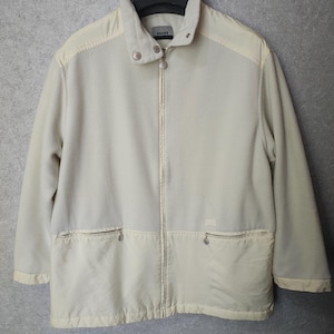 Può includere: Giacca in pile color crema con chiusura a zip completa. La giacca presenta un collo alto, due tasche frontali e un piccolo logo sul petto. La giacca è appesa a una gruccia nera.
