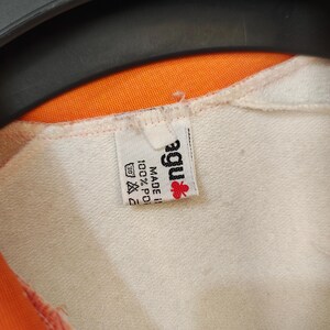 Puede incluir: Primer plano de una prenda blanca con forro naranja. Se ve una etiqueta con la marca "agu" con un logotipo de flor roja, junto con las instrucciones de lavado y la composici&oacute;n del tejido: 100% poli&eacute;ster.