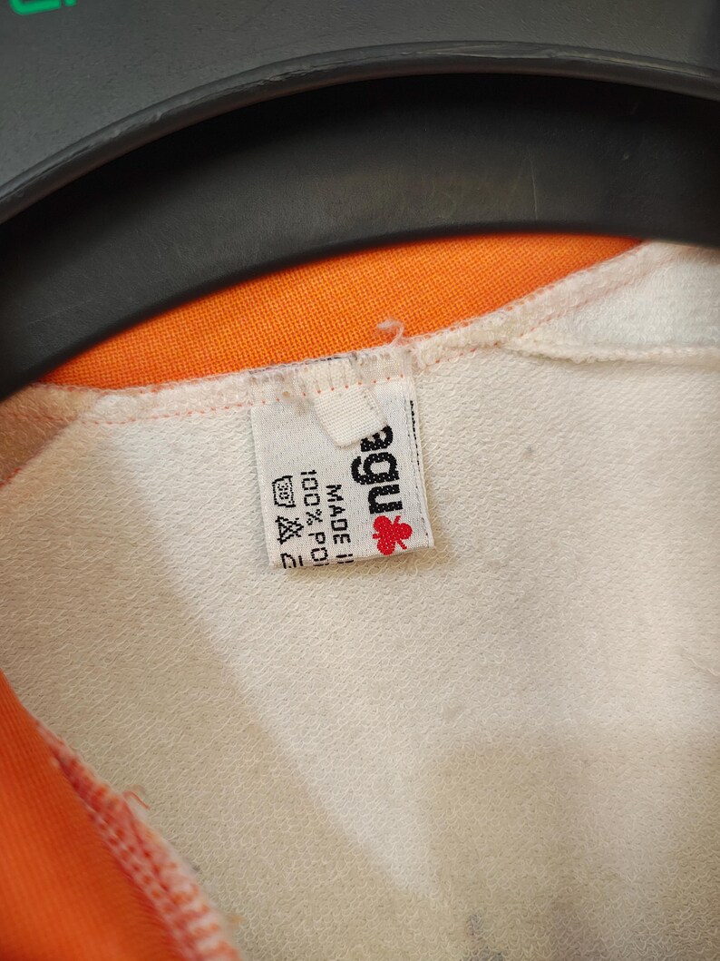 Puede incluir: Primer plano de una prenda blanca con forro naranja. Se ve una etiqueta con la marca "agu" con un logotipo de flor roja, junto con las instrucciones de lavado y la composici&oacute;n del tejido: 100% poli&eacute;ster.