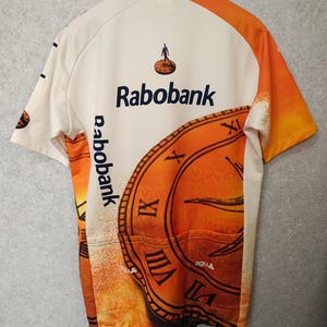 Puede incluir: Maillot de ciclismo color crema con detalles en naranja y un gran gr&aacute;fico de reloj. Presenta el logo "Rabobank" y el texto "de lage landen". Incluye tres bolsillos traseros. El maillot est&aacute; colgado de una percha negra.