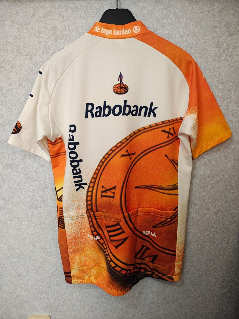 Puede incluir: Maillot de ciclismo color crema con detalles en naranja y un gran gr&aacute;fico de reloj. Presenta el logo "Rabobank" y el texto "de lage landen". Incluye tres bolsillos traseros. El maillot est&aacute; colgado de una percha negra.