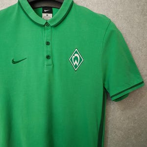Puede incluir: Polo verde con cuello y tres botones. Cuenta con un logotipo de Nike y un escudo del equipo en el pecho. La camisa tiene ribetes verde oscuro en el cuello y las mangas. El texto "cropp town" es visible en la percha.