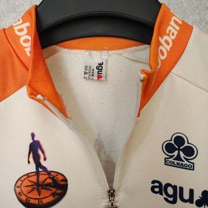 Puede incluir: Maillot de ciclismo color crema con detalles naranjas en el cuello y los hombros. Cuenta con cremallera, el logo de Colnago y la marca agu. Un gr&aacute;fico de una persona sobre una br&uacute;jula est&aacute; impreso en la parte delantera.
