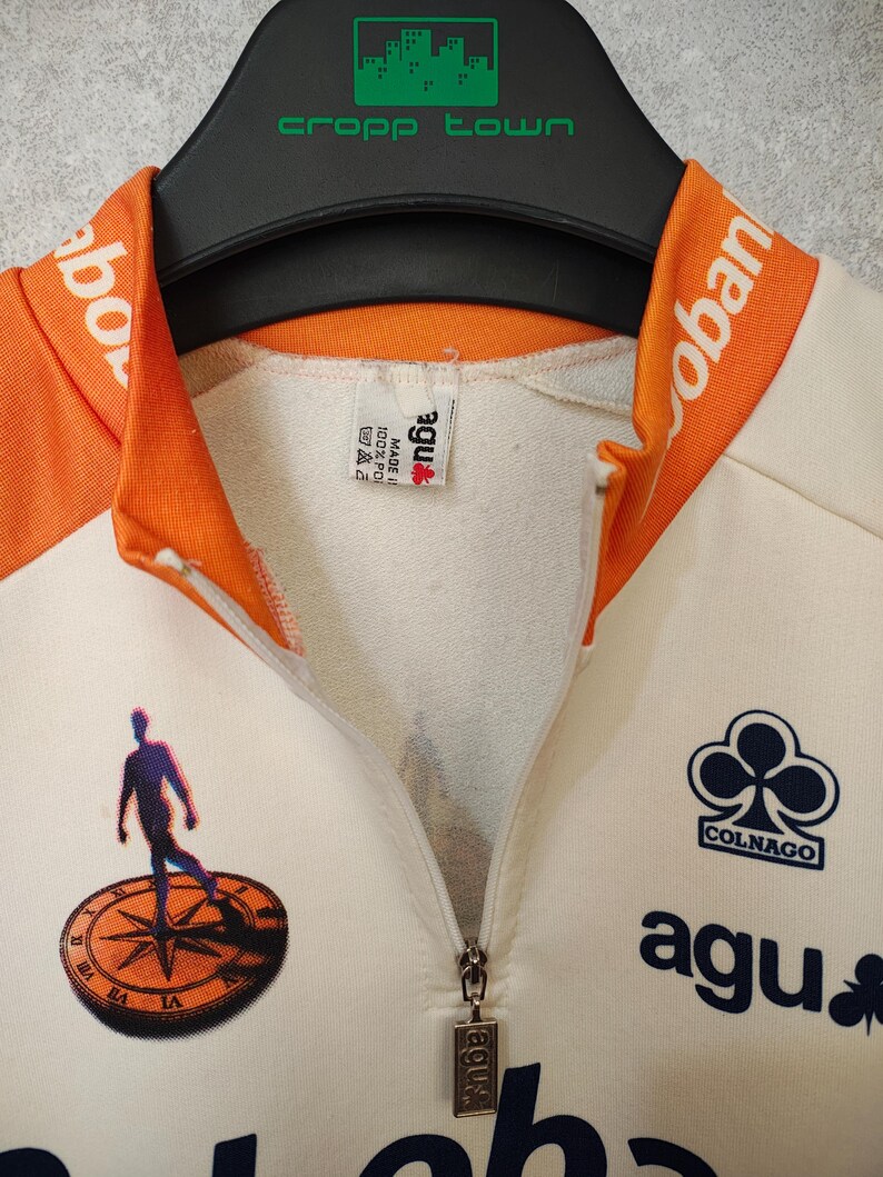 Puede incluir: Maillot de ciclismo color crema con detalles naranjas en el cuello y los hombros. Cuenta con cremallera, el logo de Colnago y la marca agu. Un gr&aacute;fico de una persona sobre una br&uacute;jula est&aacute; impreso en la parte delantera.