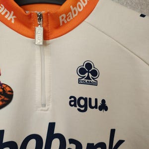 Puede incluir: Maillot de ciclismo color crema con cuello y detalles en naranja. Cuenta con cremallera, el logo "agu" y el logo "Colnago". El maillot tambi&eacute;n muestra los logos "Rabobank" y "bobank" en azul marino.