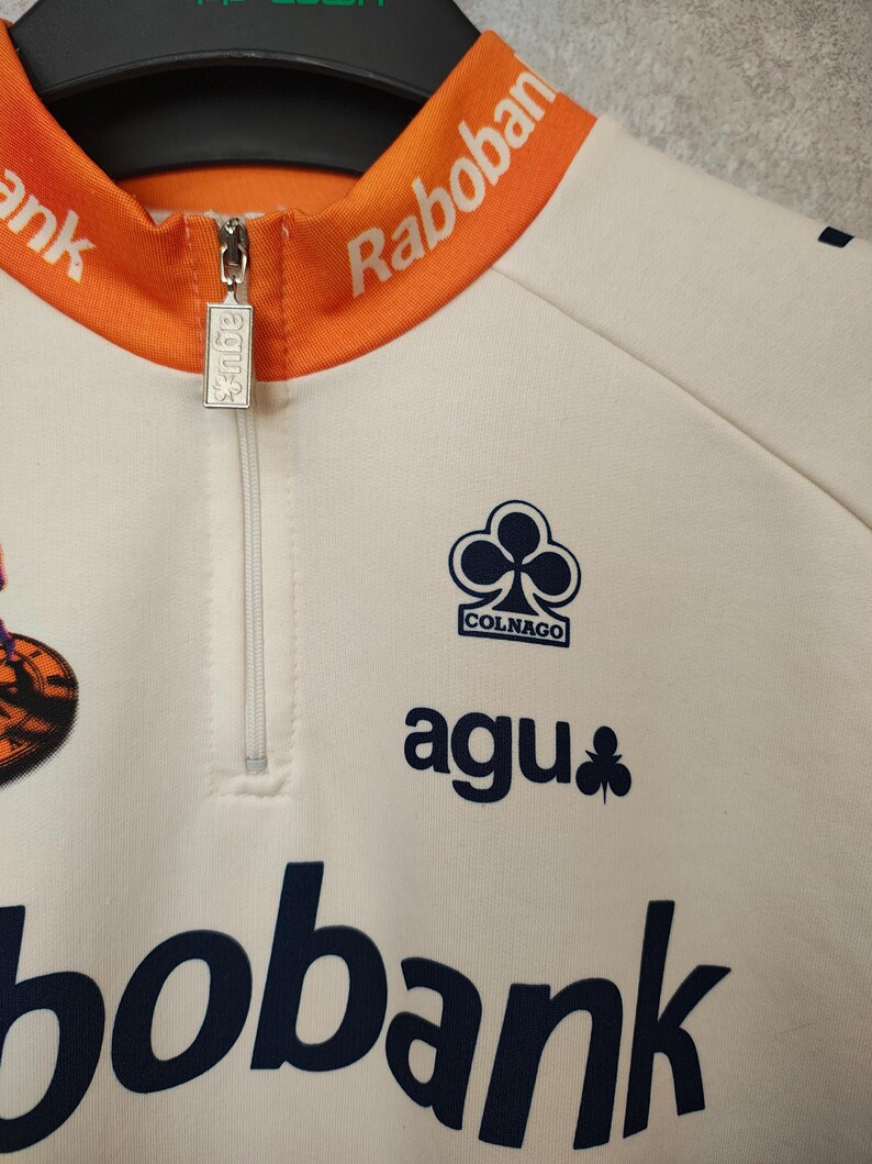 Puede incluir: Maillot de ciclismo color crema con cuello y detalles en naranja. Cuenta con cremallera, el logo "agu" y el logo "Colnago". El maillot tambi&eacute;n muestra los logos "Rabobank" y "bobank" en azul marino.