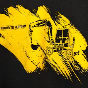K&ouml;nnte beinhalten: Schwarzes T-Shirt mit einer gelben Grafik eines Komatsu-Baggers. Der Bagger ist vor einem gelben Pinselstrich-Design platziert, mit dem Text "PROUD TO PERFORM" in Schwarz gedruckt.