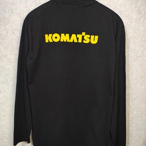 K&ouml;nnte beinhalten: Schwarzes Langarmshirt mit dem Wort "KOMATSU" in Gelb auf dem R&uuml;cken aufgedruckt. Das Shirt h&auml;ngt an einem schwarzen Kleiderb&uuml;gel vor grauem Hintergrund. Der Text ist fett und ohne Serifen.