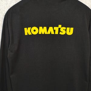 K&ouml;nnte beinhalten: Schwarzes Langarmshirt mit dem gelben Schriftzug "KOMATSU" auf dem R&uuml;cken. Das Shirt h&auml;ngt an einem schwarzen Kleiderb&uuml;gel vor grauem Hintergrund. Der Text ist fett und ohne Serifen.