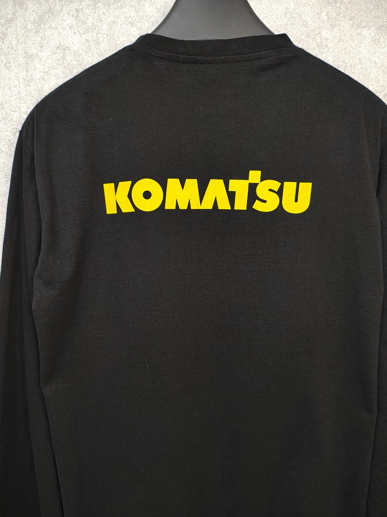 K&ouml;nnte beinhalten: Schwarzes Langarmshirt mit dem gelben Schriftzug "KOMATSU" auf dem R&uuml;cken. Das Shirt h&auml;ngt an einem schwarzen Kleiderb&uuml;gel vor grauem Hintergrund. Der Text ist fett und ohne Serifen.