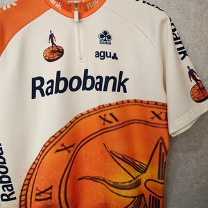 Puede incluir: Maillot de ciclismo color crema con detalles en naranja y el logo "Rabobank". Cuenta con cremallera parcial, dise&ntilde;o de reloj y el logo "Colnago agu". El maillot tiene mangas cortas y un peque&ntilde;o gr&aacute;fico de un ciclista.