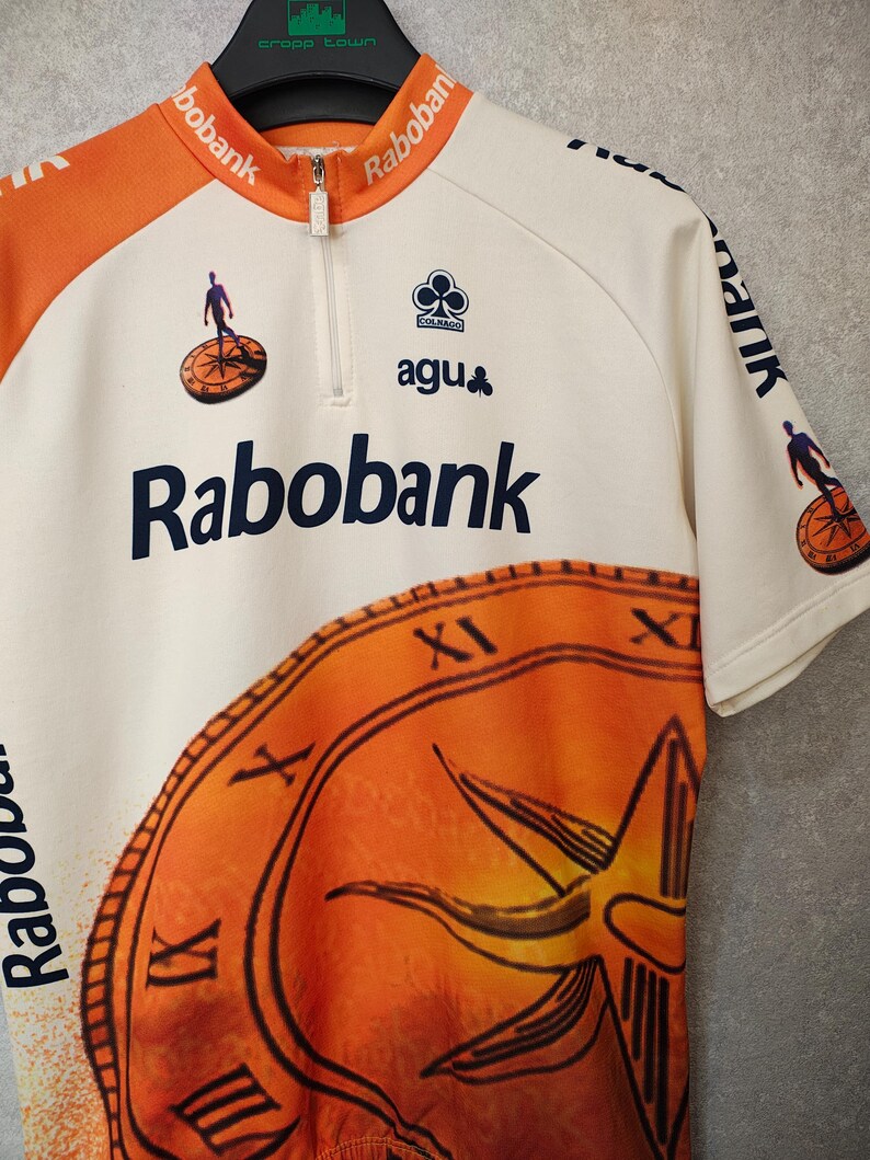 Puede incluir: Maillot de ciclismo color crema con detalles en naranja y el logo "Rabobank". Cuenta con cremallera parcial, dise&ntilde;o de reloj y el logo "Colnago agu". El maillot tiene mangas cortas y un peque&ntilde;o gr&aacute;fico de un ciclista.