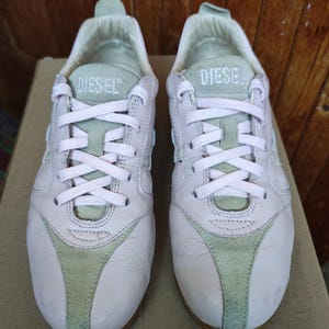 Peut inclure: Une paire de baskets rose clair et vert pâle avec une fermeture à lacets et le mot DIESEL imprimé sur la languette. Les chaussures sont fabriquées avec une combinaison de panneaux en cuir lisse et en daim, incluant une tirette au talon.