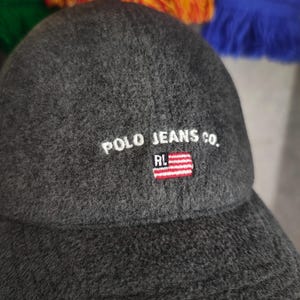 May include: A dark gray baseball cap with the text "POLO JEANS CO." embroidered in white. The cap features a small American flag patch with red and white stripes and a blue field with white stars. The cap is made of a textured fabric.