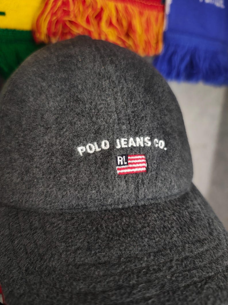May include: A dark gray baseball cap with the text "POLO JEANS CO." embroidered in white. The cap features a small American flag patch with red and white stripes and a blue field with white stars. The cap is made of a textured fabric.