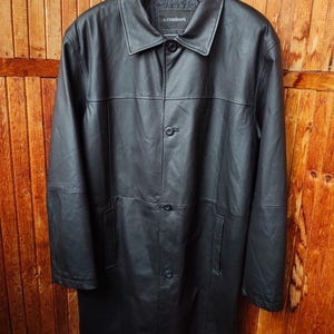 May include: A black leather coat with a button-up front and a collared neckline. The coat is hanging on a black hanger against a wooden background. The brand name "c.comberti" is visible on the label.