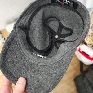 May include: A gray fleece cap with a curved brim. The interior features a black lining and a star-shaped support structure. The label reads "POLO JEANS CO. RALPH LAUREN."