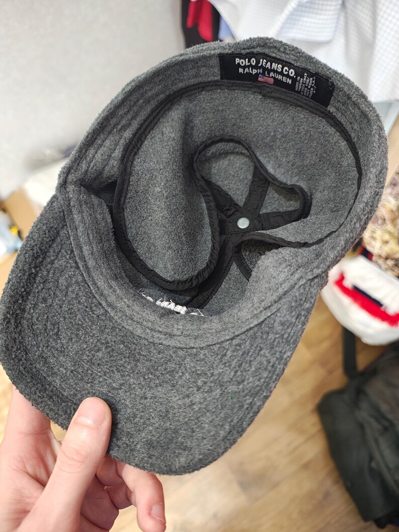 May include: A gray fleece cap with a curved brim. The interior features a black lining and a star-shaped support structure. The label reads "POLO JEANS CO. RALPH LAUREN."
