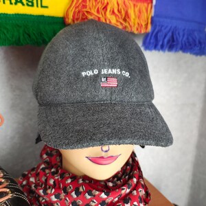 May include: A gray Polo Jeans Co. baseball cap with the brand name and logo embroidered on the front. The cap has a curved brim and is displayed on a mannequin head. A red patterned scarf is wrapped around the mannequin's neck.