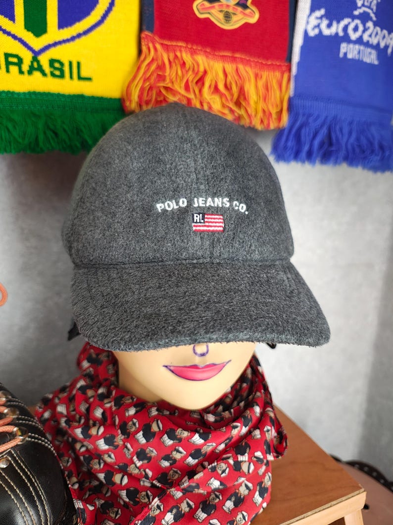 May include: A gray Polo Jeans Co. baseball cap with the brand name and logo embroidered on the front. The cap has a curved brim and is displayed on a mannequin head. A red patterned scarf is wrapped around the mannequin's neck.