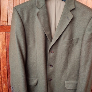 May include: A brown tweed blazer with a classic three-button closure, notched lapels, and two front pockets. The blazer is hanging on a black hanger against a wooden background. The blazer has a subtle herringbone pattern.