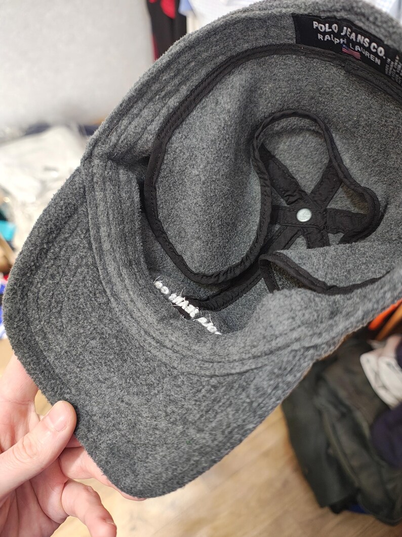 May include: A gray Polo Jeans Co. baseball cap with a curved brim. The interior features a black lining and a white label with text. The cap is made of a soft, textured material.