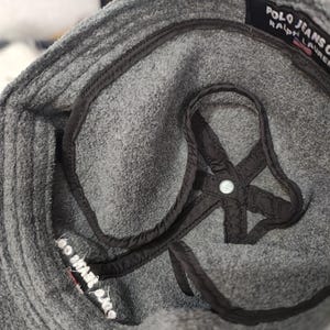 May include: Close-up of a gray Polo Jeans Co. hat. The hat has a black interior lining and a black strap detail. The brand name is visible on a black tag. The hat is made of a soft, textured material.
