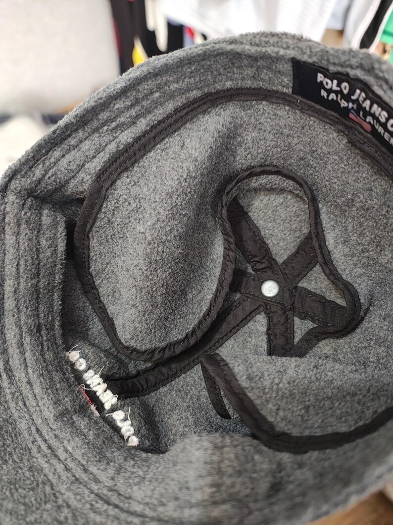 May include: Close-up of a gray Polo Jeans Co. hat. The hat has a black interior lining and a black strap detail. The brand name is visible on a black tag. The hat is made of a soft, textured material.
