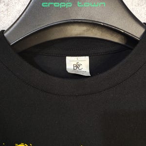 K&ouml;nnte beinhalten: Schwarzes T-Shirt mit Rundhalsausschnitt und gelbem Pinselstrich-Design. Das Shirt h&auml;ngt an einem schwarzen Kleiderb&uuml;gel mit dem gr&uuml;nen Text "cropp town". Ein Etikett mit den Buchstaben "B&C COLLECTION" ist sichtbar.