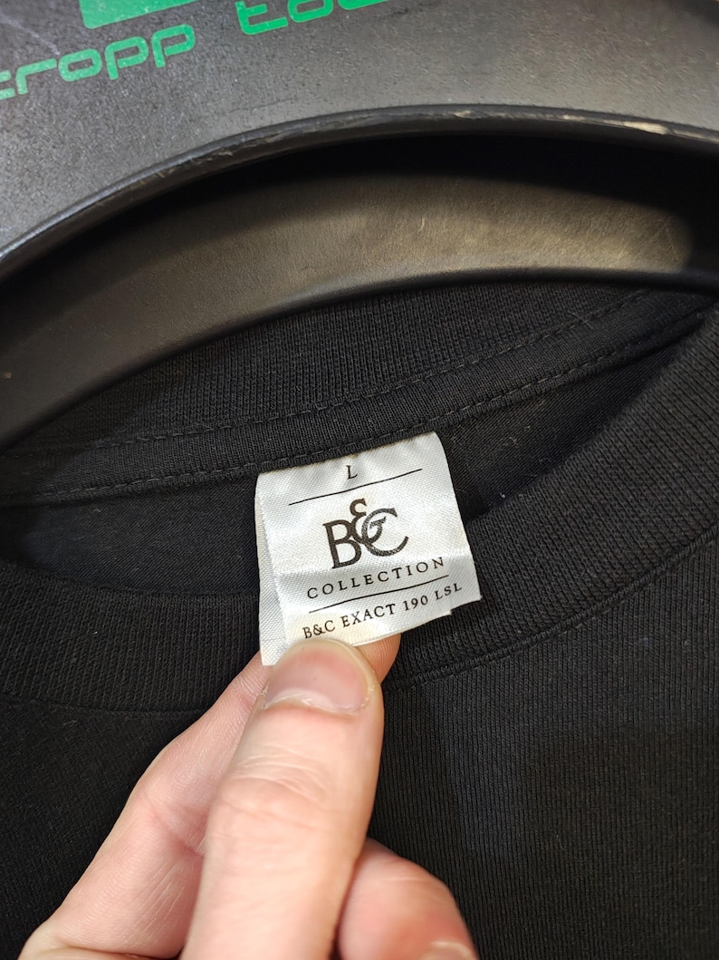 K&ouml;nnte beinhalten: Ein schwarzes T-Shirt mit einem wei&szlig;en Etikett. Auf dem Etikett steht die Gr&ouml;&szlig;e "L", sowie die Aufschrift "B&C COLLECTION" und "B&C EXACT 190 LSL". Das T-Shirt wird von einer Person gehalten.