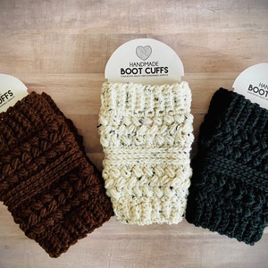 May include: Three pairs of handmade crocheted boot cuffs in brown, white, and black. Each cuff has a tag that reads "Handmade Boot Cuffs".