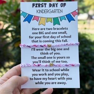 May include: Two braided bracelets with pink, purple, and white threads, each featuring a small gold-colored heart charm. They are attached to a white card with colorful bunting graphics and text reading First Day of Kindergarten and a poem.