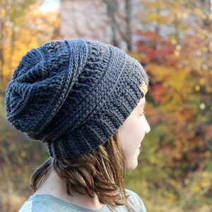 May include: A person wearing a gray crocheted beanie with a brown leather label. The beanie is slouchy and has a textured design. The person is looking off to the side and has long brown hair.
