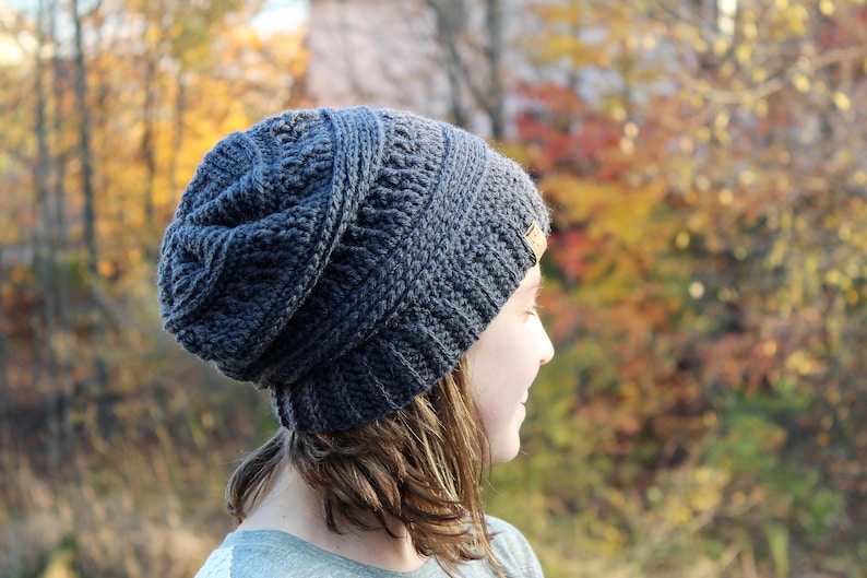May include: A person wearing a gray crocheted beanie with a brown leather label. The beanie is slouchy and has a textured design. The person is looking off to the side and has long brown hair.