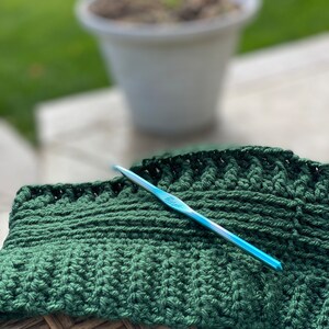 May include: A close-up of a green crocheted blanket with a blue crochet hook resting on top. The blanket is sitting on a woven wicker surface.