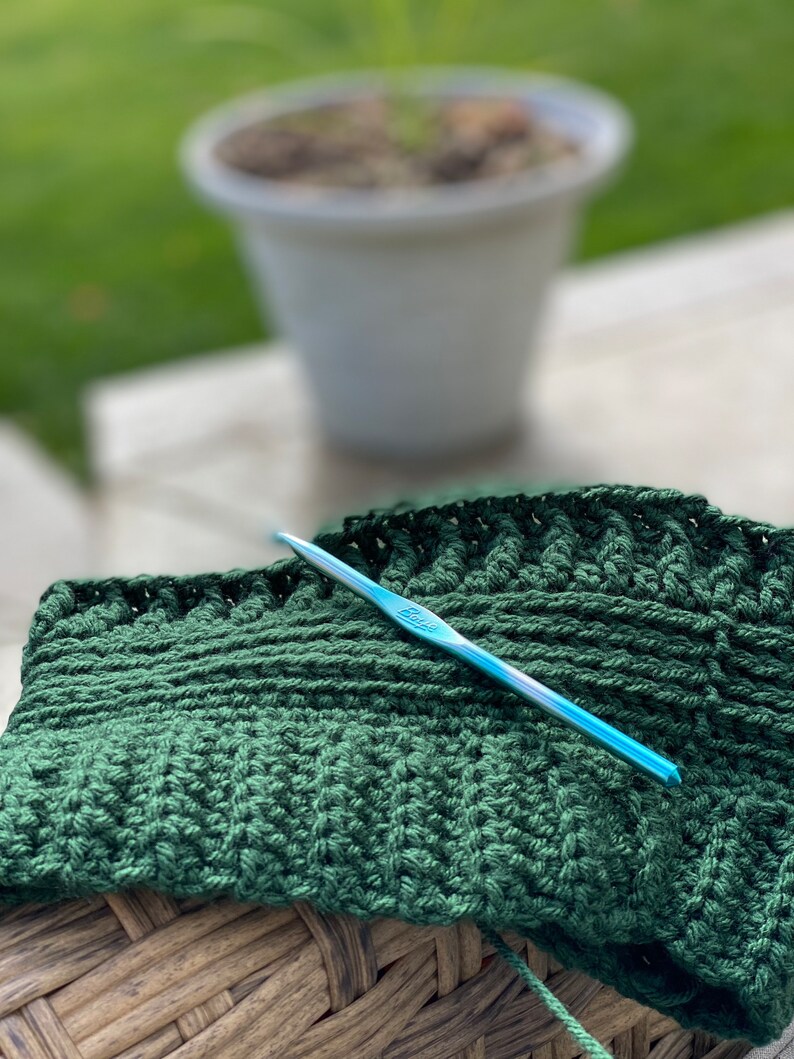 May include: A close-up of a green crocheted blanket with a blue crochet hook resting on top. The blanket is sitting on a woven wicker surface.