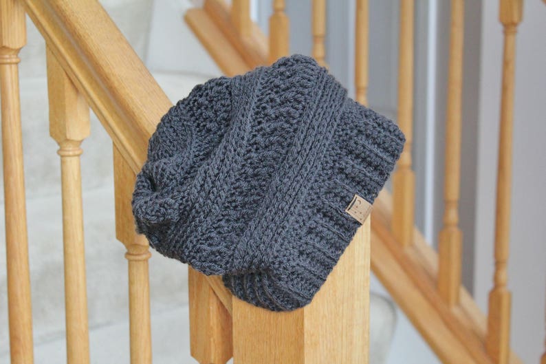 May include: A gray knitted beanie with a textured pattern. The beanie has a small brown label attached to the side.