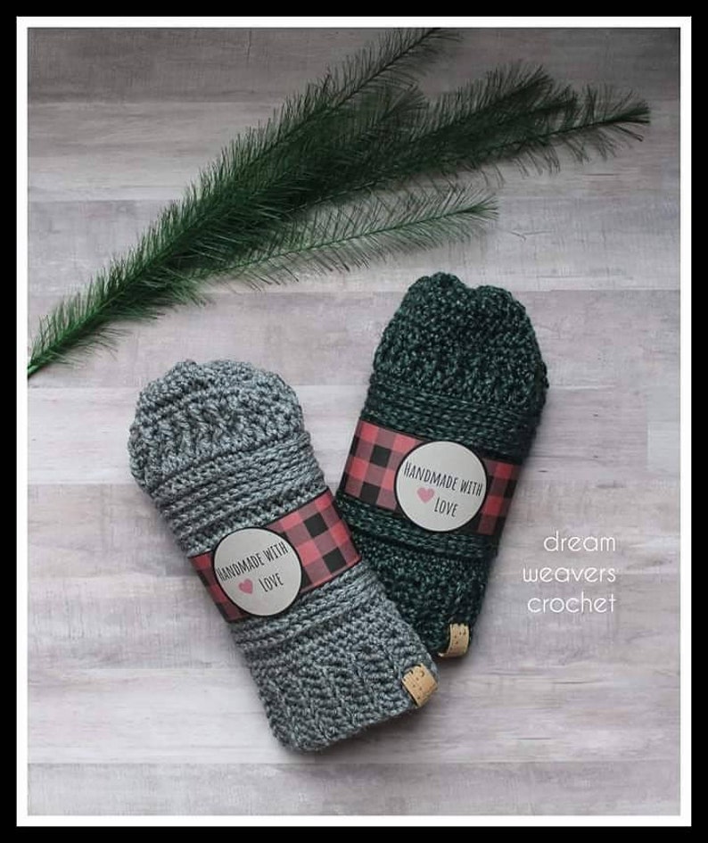 May include: Two crocheted fingerless gloves, one gray and one green, with a red and black buffalo plaid label that says "Handmade with Love". The gloves are on a light wood surface. The text "dream weavers crochet" is written below the gloves.