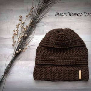 May include: A brown crocheted beanie with a textured stitch pattern. The beanie has a fold-over cuff and a brown leather tag with the text "Dream Weavers Crochet".