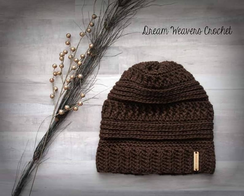 May include: A brown crocheted beanie with a textured stitch pattern. The beanie has a fold-over cuff and a brown leather tag with the text "Dream Weavers Crochet".