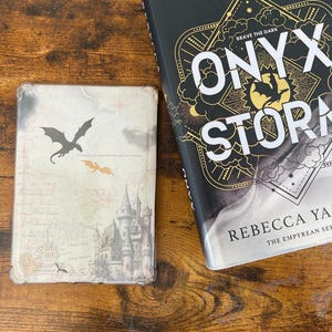 May include: Two books on a wooden surface. The larger book, "Onyx Storm" by Rebecca Yarros, has a black cover with gold accents and white text. The smaller book has a dragon graphic and a castle illustration. The text on the larger book reads "Brave the Dark."