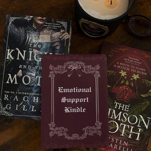 May include: A dark maroon rectangular Kindle skin featuring a white ornate border with a bow at the top and the text Emotional Support Kindle in the centre, positioned between two books and next to a lit candle in a black container.