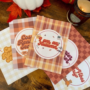 Könnte beinhalten: Vier rechteckige Karten mit Karomustern in Orange-, Braun- und Cremetönen. Jede Karte zeigt ein kreisförmiges Emblem mit Text, darunter "Cozy Reading Season", "I am an Autumn", "Sweater Weather" und "Cozy Vibes" sowie kleinen Symbolen.